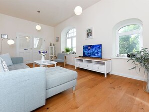 A living room with a light blue sofa, a white entertainment unit, and a flat-screen TV. There are two windows with arched tops, a door, and a potted plant.