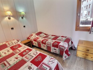 Une chambre avec deux lits, chacun recouvert d’une couette rouge et blanche, une commode en bois et deux lampes fixées au mur.