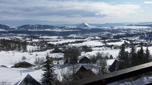 A panoramic view of a snow-covered landscape with mountains in the distance, a few houses, and a clear sky.