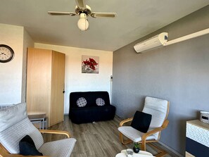 A modern living room with a ceiling fan, a clock, a black sofa with patterned cushions, two armchairs, a small table with a glass, and a wall-mounted air conditioning unit.