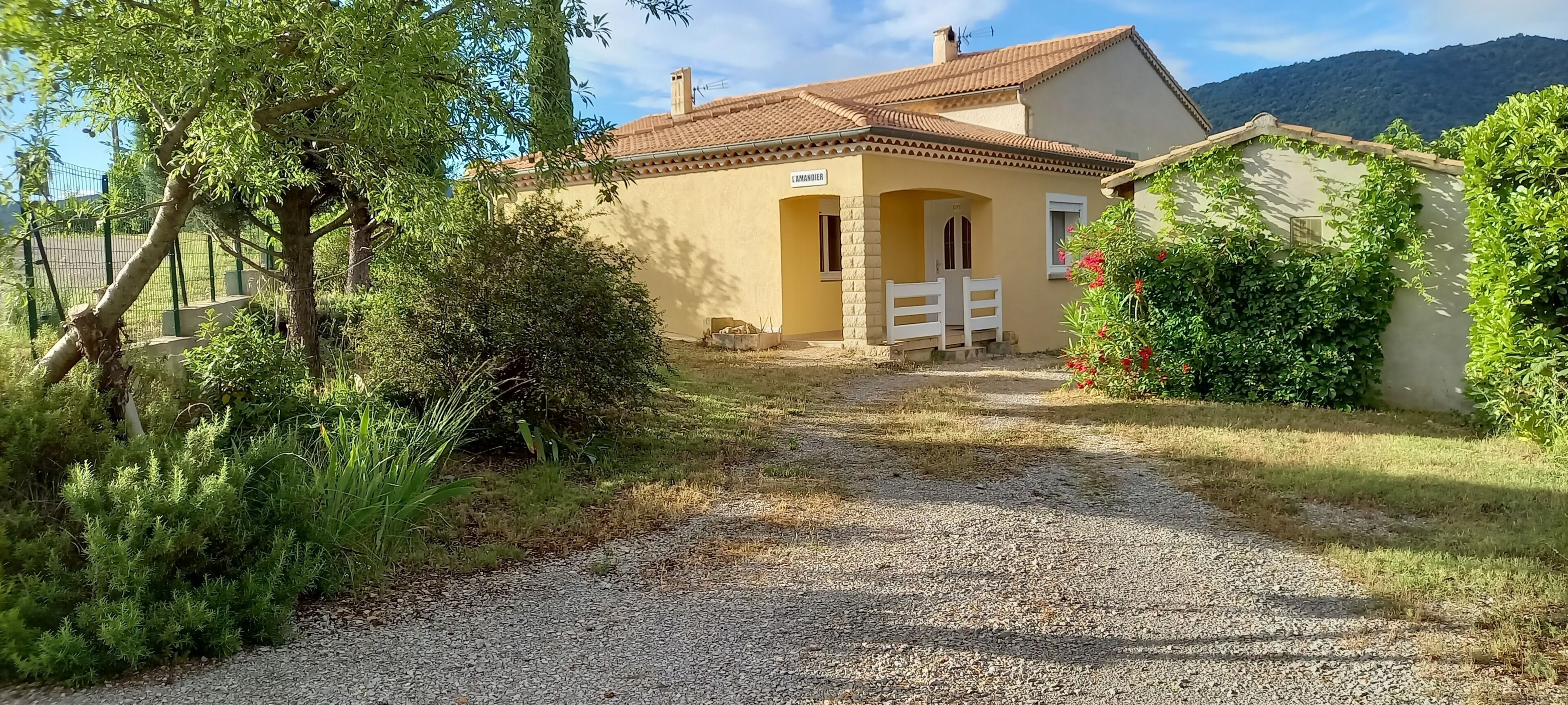 Una casa con vialetto sterrato, cancello in legno e una montagna sullo sfondo.