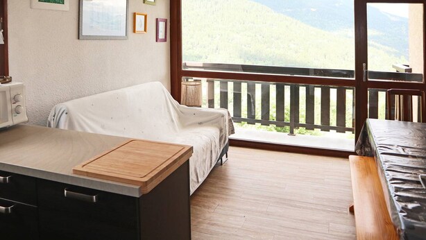A cozy room with a bed, a wooden table, a kitchenette, and a balcony with a mountain view.