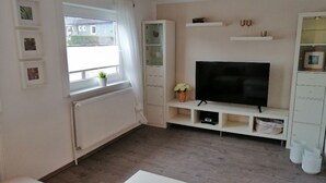 A living room with a large flat-screen TV, white cabinetry, a radiator, and a window with a view of the outside. - Vacation apartment/holiday home for up to 5 people (children under 6 free of charge) (Clausthal-Zellerfeld)