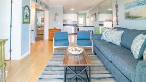 Uma sala de estar com sofá, duas poltronas, uma mesa de centro e uma cozinha ao fundo.