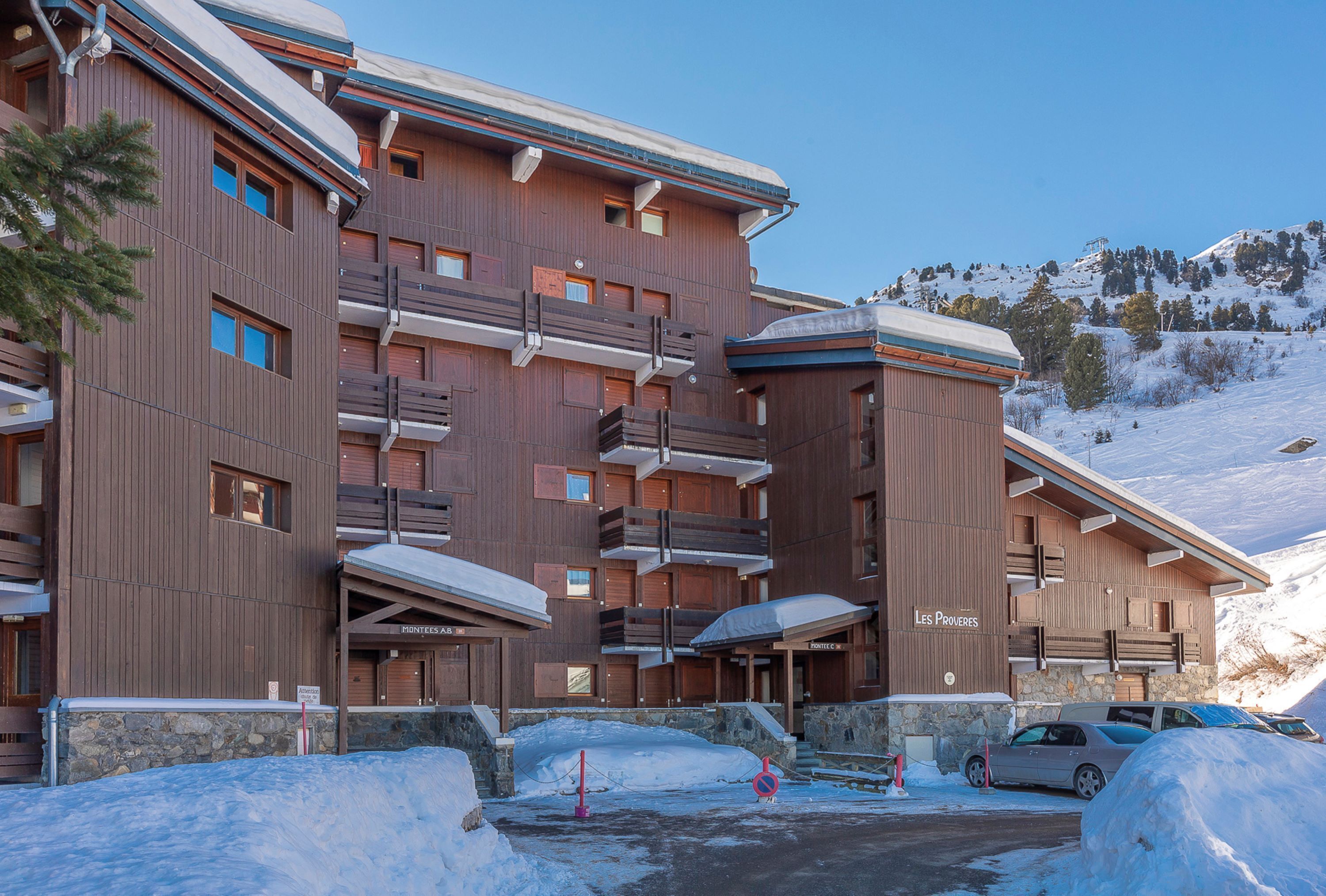A multi-story wooden building with balconies, a ski slope in the background, and a sign reading "Les Proveres."