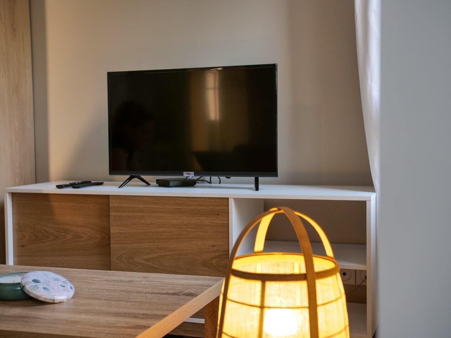 Una sala moderna con una televisión de pantalla plana sobre un pedestal blanco, una mesa de madera con una lámpara y un mueble debajo.