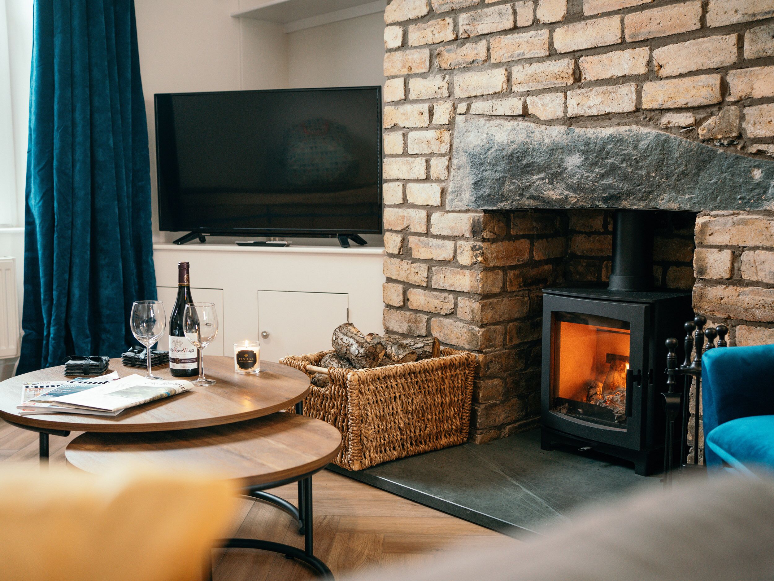 A cozy living room with a brick fireplace, a flat-screen TV mounted on the wall, and a round wooden table with magazines and a bottle of wine.