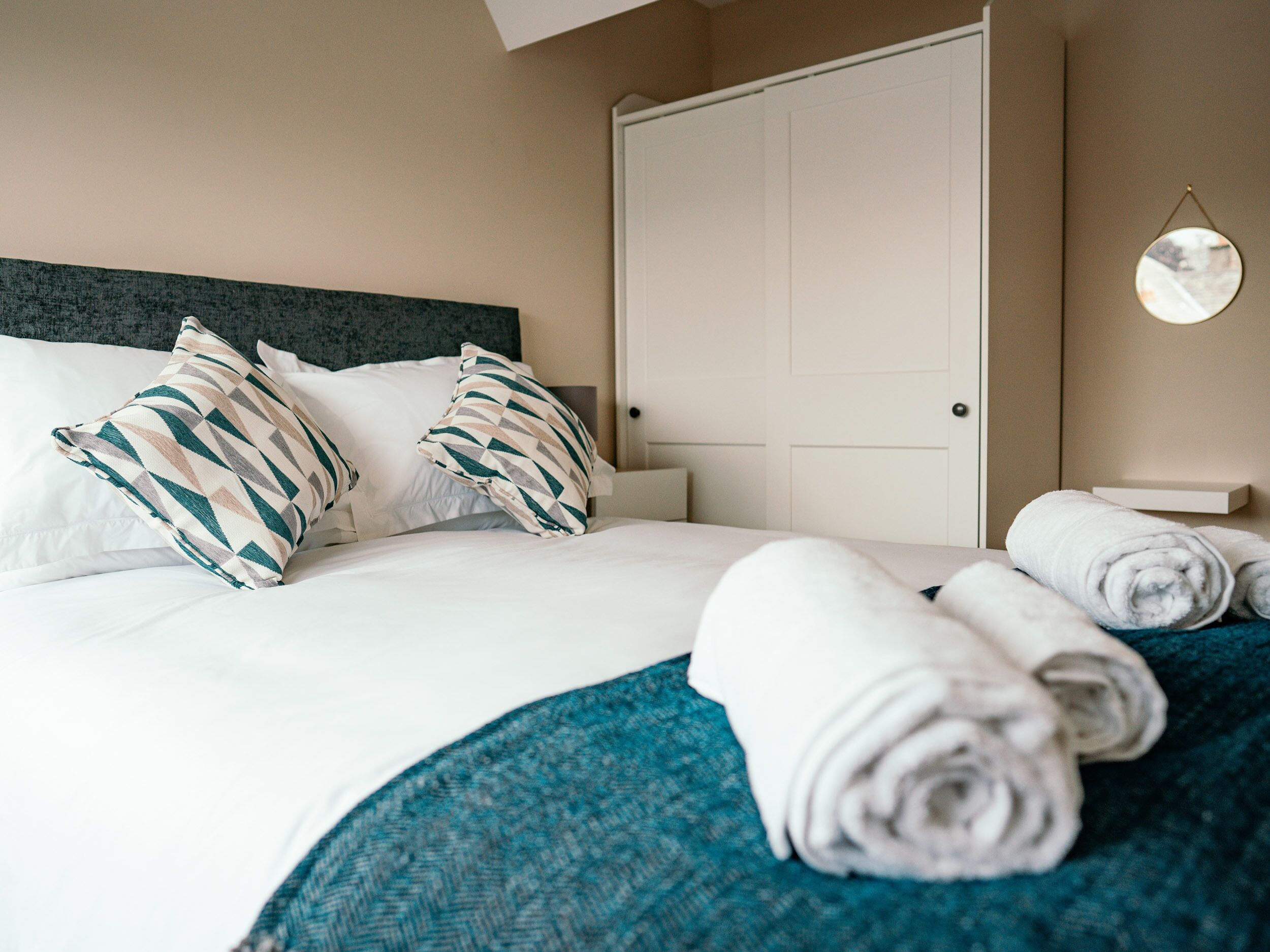 A neatly made bed with white linens and patterned pillows, a teal throw blanket, and rolled towels on the side.