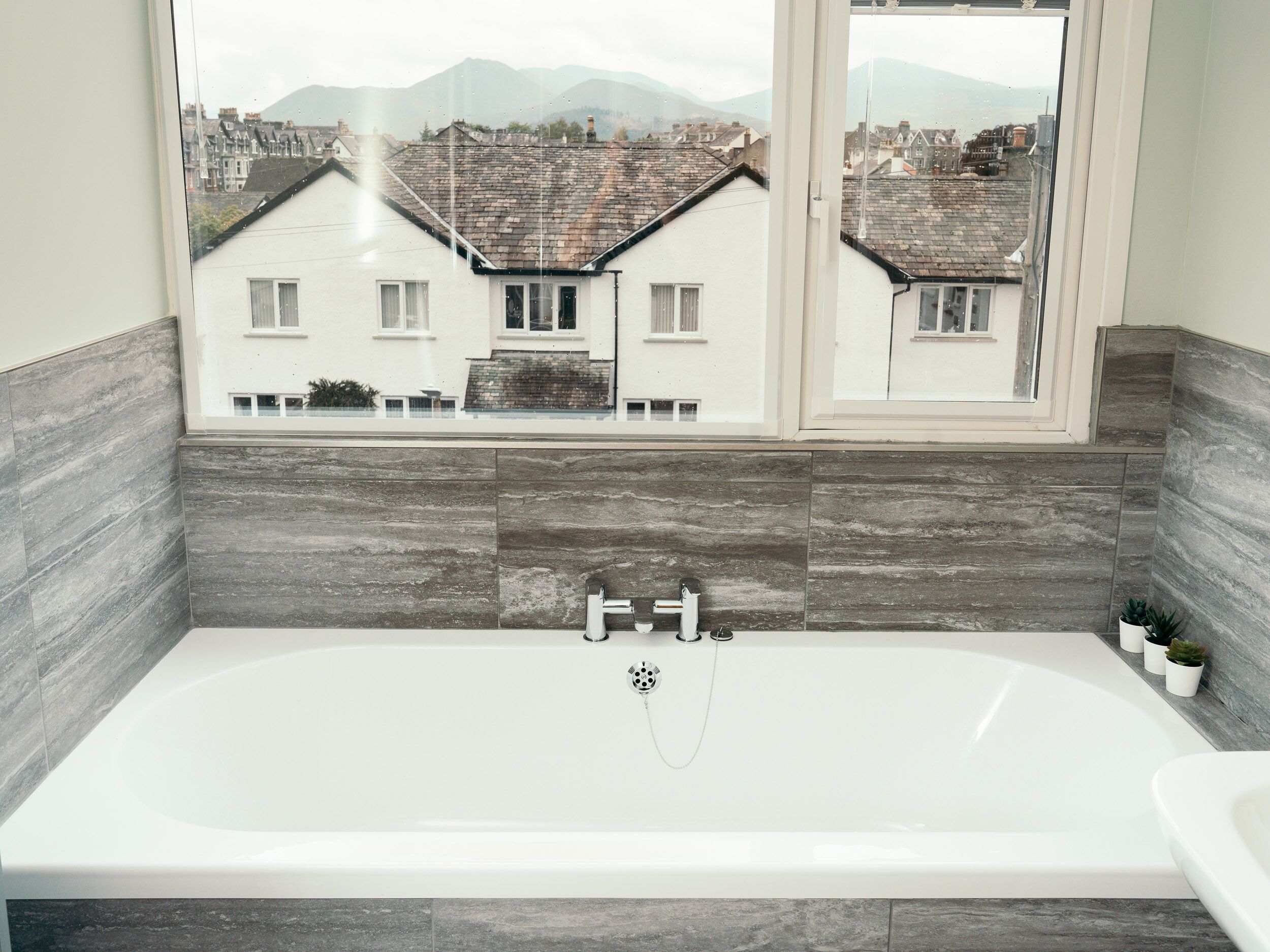 A modern bathroom with a bathtub, a window with a view of houses and mountains, and grey tiled walls.