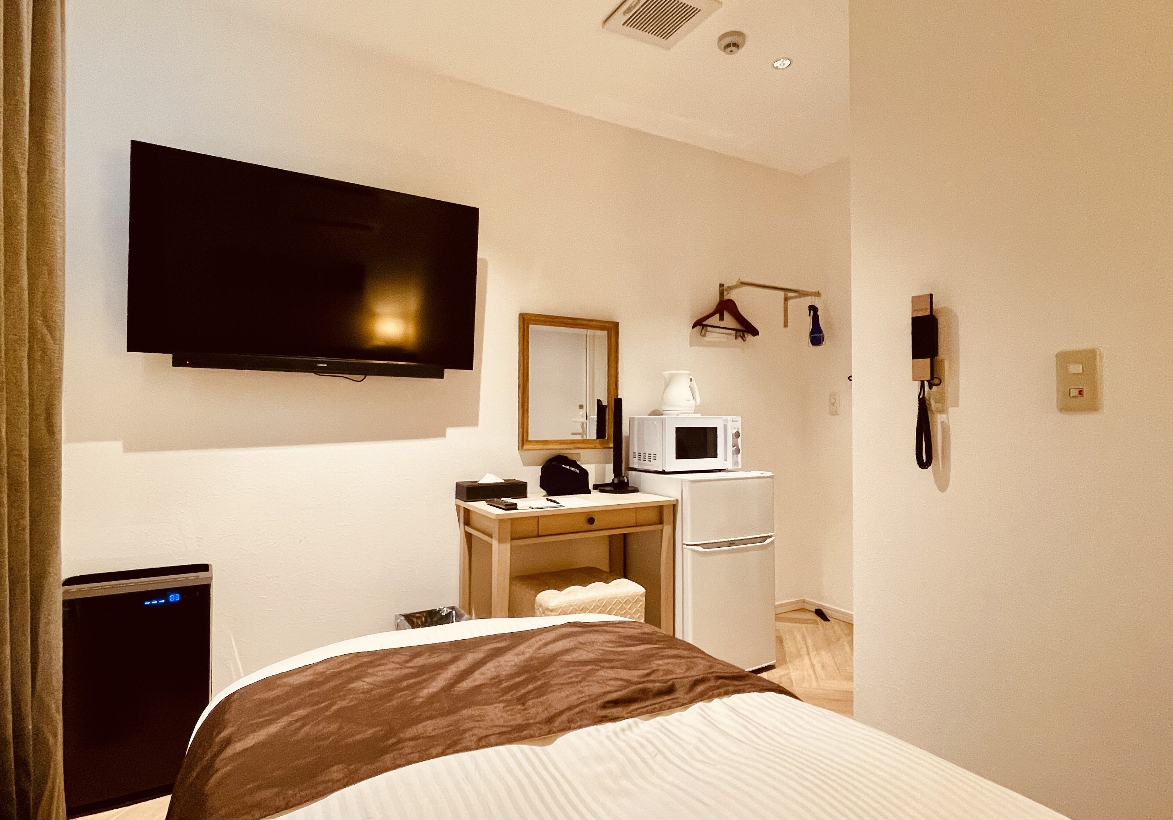 A hotel room with a bed, a desk with a computer, a microwave, a refrigerator, a TV, and a telephone.
