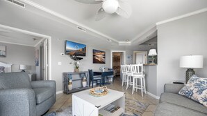 Un salon comprenant un canapé, une table basse, un téléviseur, un coin repas avec des chaises et un ventilateur de plafond.