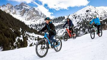 Tres ciclistas de montaña en un sendero nevado, con montañas cubiertas de nieve al fondo.