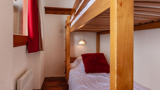 Une petite pièce avec un lit superposé, une fenêtre ornée de rideaux rouges et un luminaire fixé au mur.