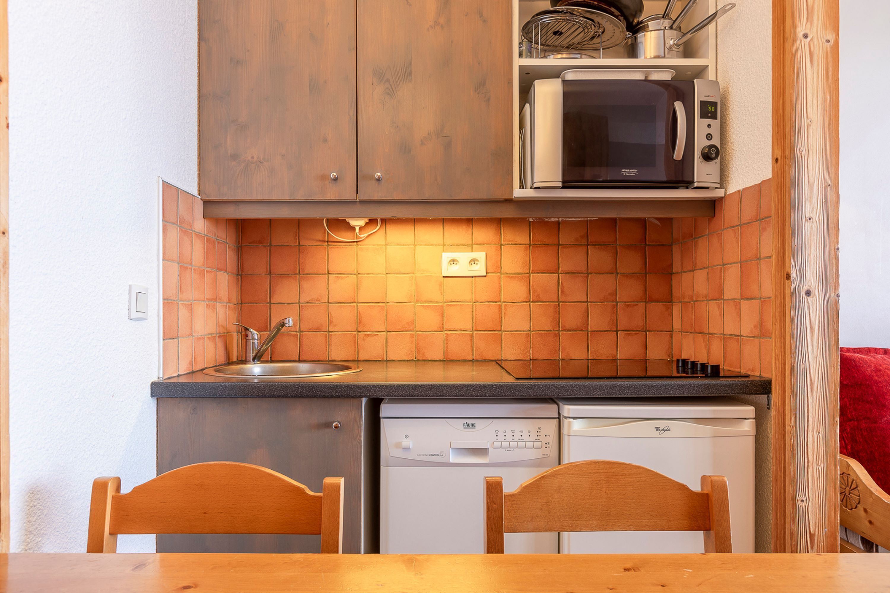 A compact kitchen with wooden cabinets, a microwave, a dishwasher, and two wooden chairs at a table.