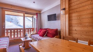 A cozy room with a wooden table, a sofa with red cushions, and a TV mounted on the wall. - Résidence Le Pralin - Méribel-Mottaret (Les Allues)
