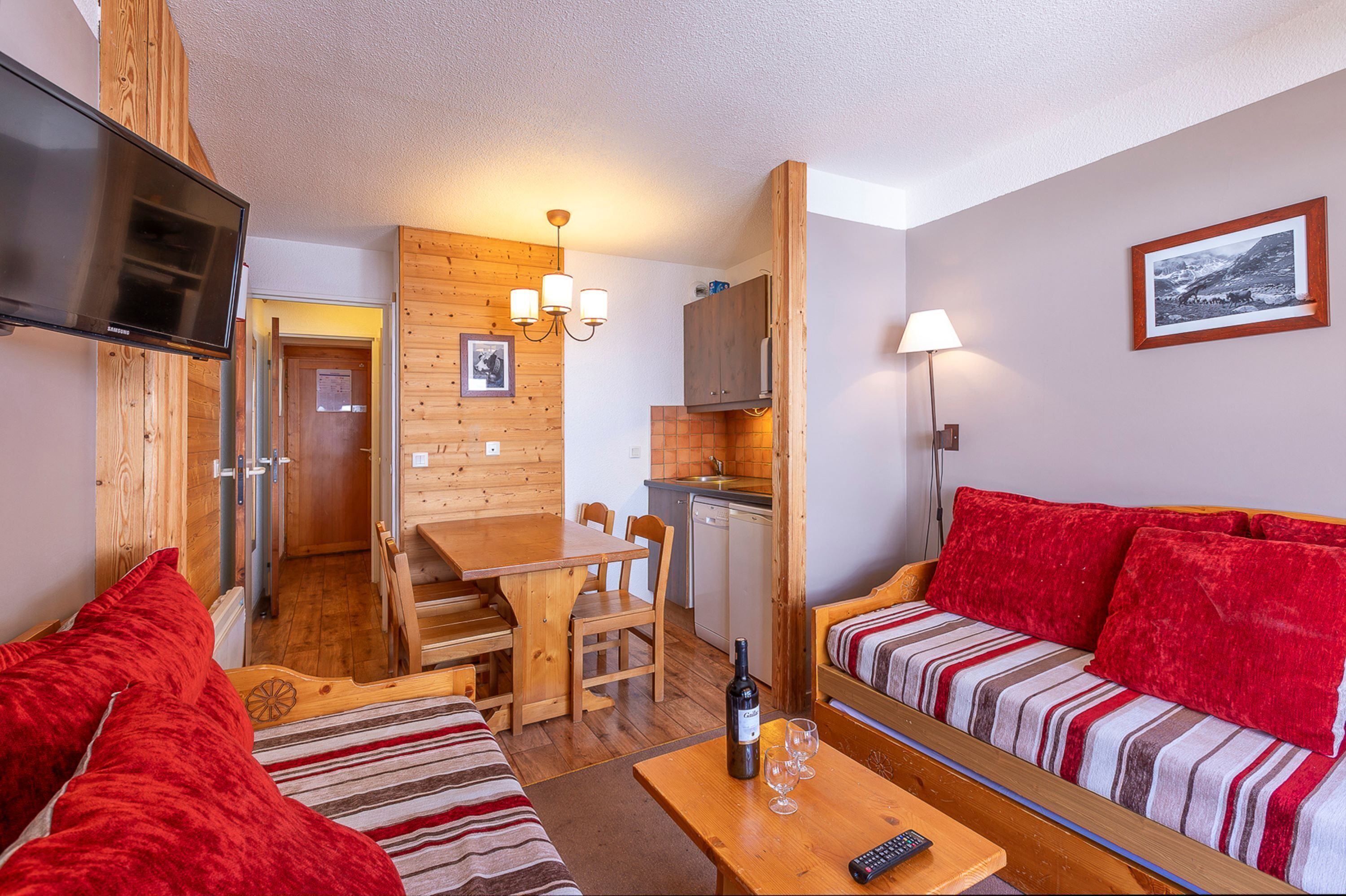 A cozy room with a wooden dining table, a sofa with red cushions, a TV, and a kitchen area with a sink and cabinets.