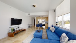 A modern living room with a large flat-screen TV, a blue sofa, and a dining area with a kitchen in the background.