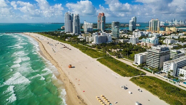 An aerial view of a coastal city with a beach, high-rise buildings, and a clear blue sky.