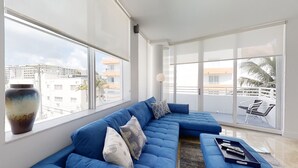 A modern living room with a blue sectional sofa, a large window offering a view of buildings and palm trees, and a balcony with chairs.