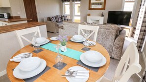 Un coin repas avec une table ronde en bois, dressée pour quatre personnes, entourée de chaises blanches. On aperçoit un canapé et un téléviseur à l’arrière-plan.