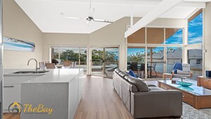 A spacious kitchen with a central island, a dining area with a table and chairs, a sofa, and a large sliding glass door leading to an outdoor patio. - Moy at Nelson Bay - 2/30 Thurlow Avenue (Nelson Bay)