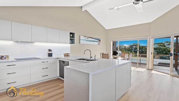 A modern kitchen with white cabinetry, a central island, and a view of the outdoors through large sliding glass doors. - Moy at Nelson Bay - 2/30 Thurlow Avenue (Nelson Bay)