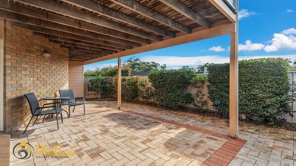 A covered patio area with brick flooring, a table and chairs, and a view of a garden and sky.