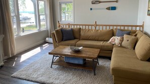 A cozy living room with a beige sofa, a wooden coffee table, and two windows with curtains. - Maryview Beach House, Maryview Beach House (La Grand Township)