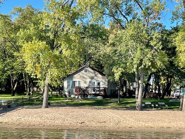 A cabin surrounded by trees, with a sandy area in front and a body of water nearby.