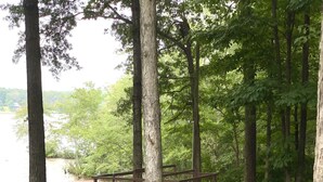 Une terrasse en bois avec des chaises et un petit bateau, entourée d’arbres et d’une zone herbeuse.