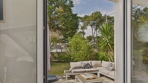 Een open glazen schuindeur die uitkomt op een terras met een beige loungeset, een houten bijzettafeltje en een vloer van stenen tegels.