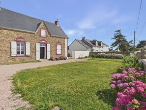 A stone house with a paved driveway, a grassy lawn, and a flower bed with pink flowers.