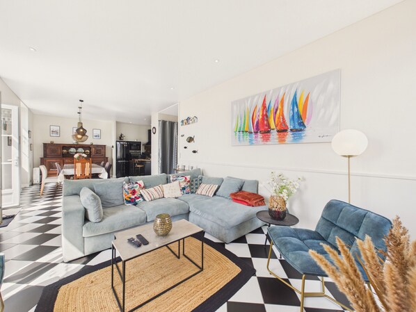 A modern living room with a checkered floor, a sectional sofa, a blue armchair, a coffee table, and a painting of sailboats.