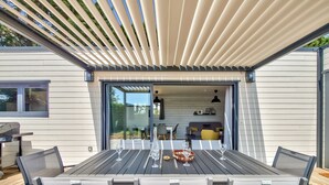 Ein überdachter Essbereich im Freien mit einem Tisch für vier Personen, eine Glastür zu einem Innenraum und einer Pergola darüber.