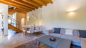 Ein modernes Wohnzimmer mit Holzbalkendecke, einem grauen Sofa, einem Couchtisch und einem Essbereich mit Stühlen und einem Tisch.