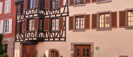 Un bâtiment traditionnel à colombages, doté de nombreuses fenêtres et portes, une rue pavée et de petits pots de plantes à l’entrée.