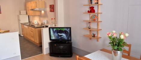 A compact living space with a kitchenette, a flat-screen TV, a wooden shelf with books and a teddy bear, and a dining area with a table and chairs.