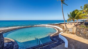 A circular pool with a stone wall and metal railing, surrounded by a sandy beach and palm trees, overlooking the ocean.
