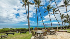 An outdoor patio with a table and chairs, surrounded by palm trees and a view of the ocean. - Beachfront Wailea condo w/ direct beach access and pools (Wailea)