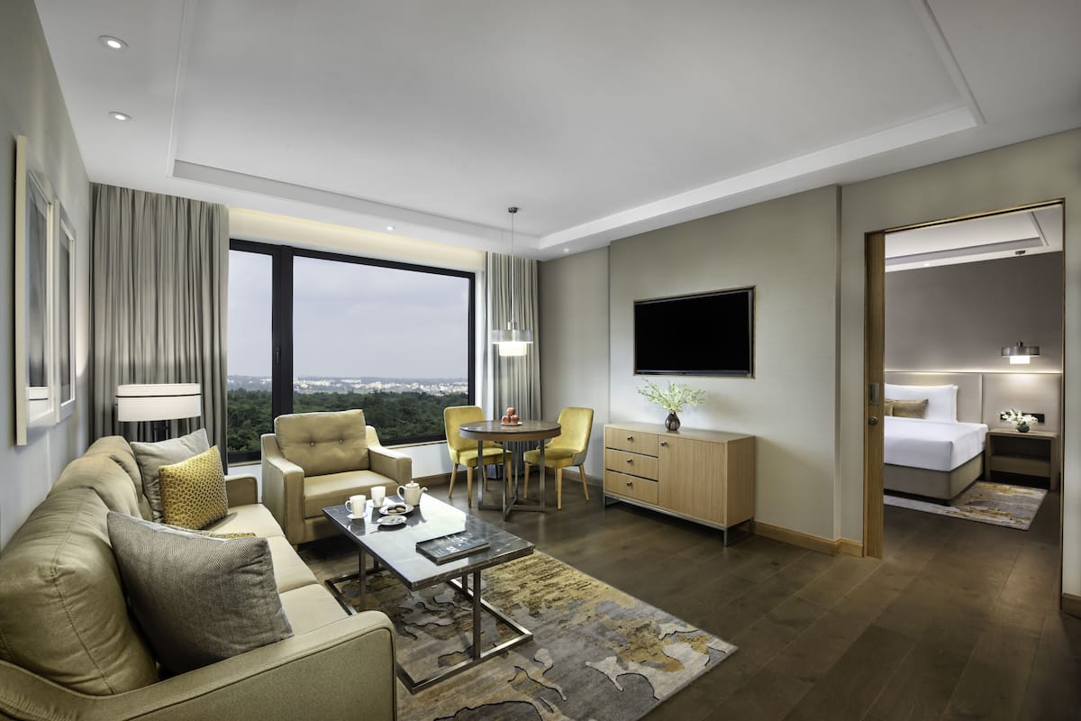 Ett modernt hotellrum med en soffa, ett matbord, stolar, en TV, ett sängbord med en lampa och utsikt över staden.