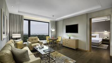 A modern hotel room with a sofa, dining table, chairs, a TV, a bedside table with a lamp, and a view of the city.