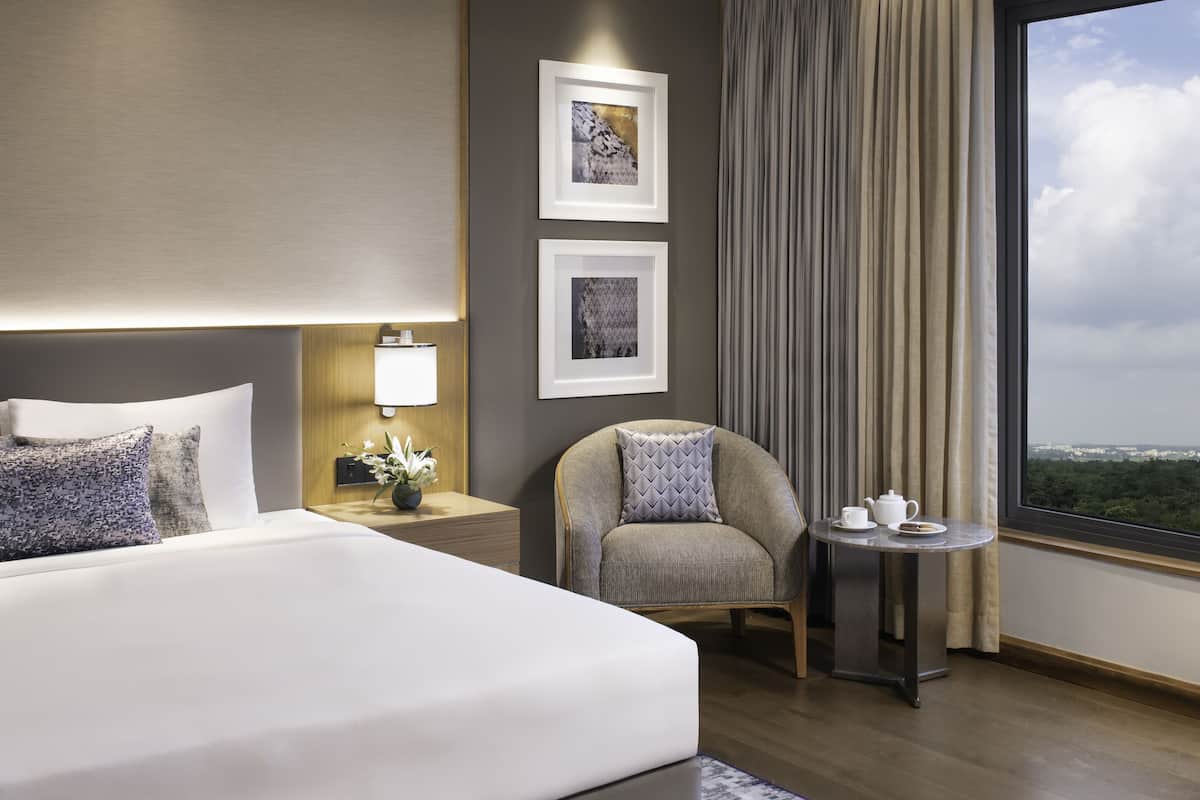 Ett modernt hotellrum med en stor säng, en stol, ett litet bord med en tekanna och utsikt över staden genom fönstret.