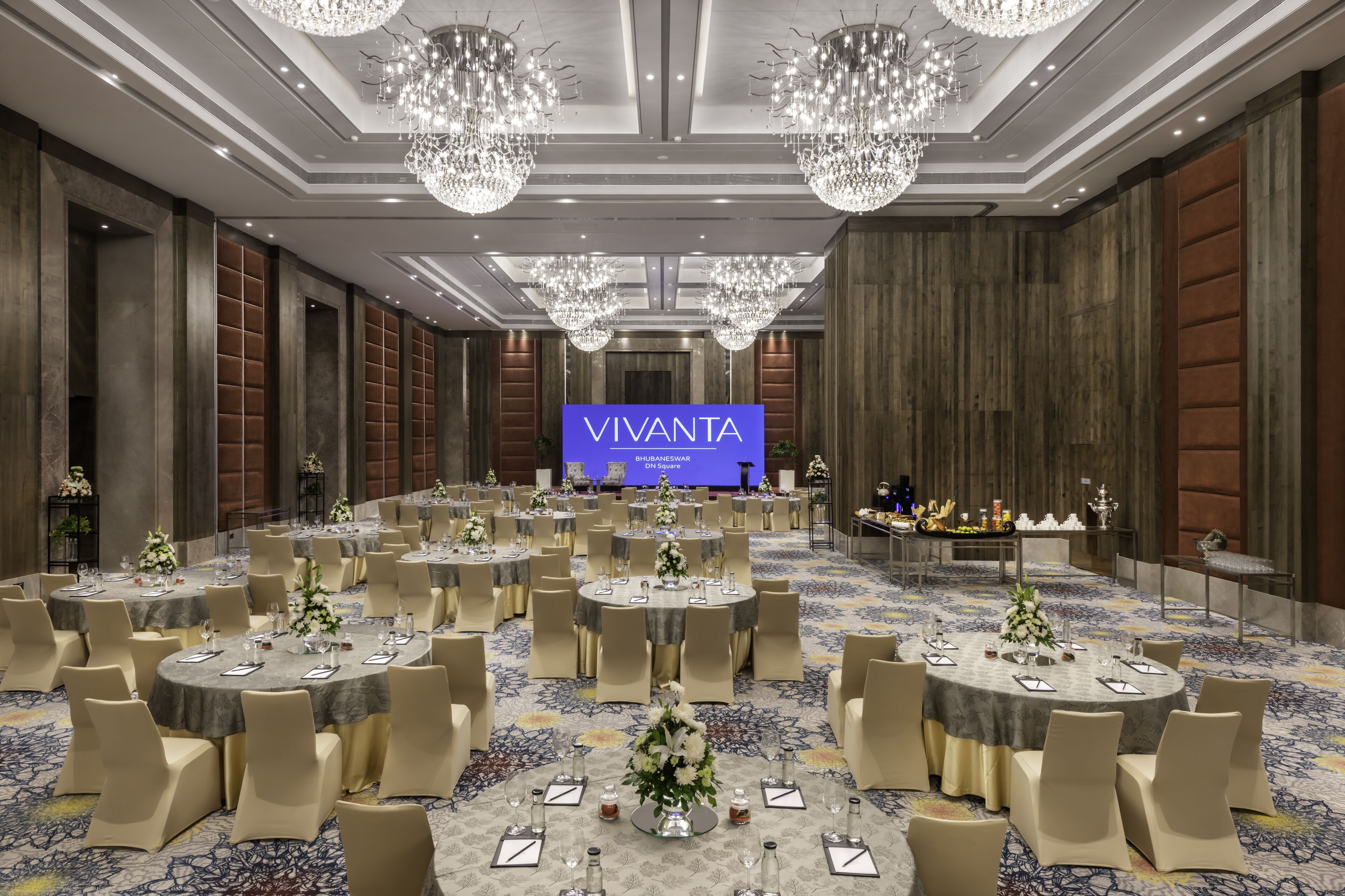 A large conference room with round tables, beige chairs, and chandeliers. A large screen displays the name "VIVANTA" and "Bhubaneswar".