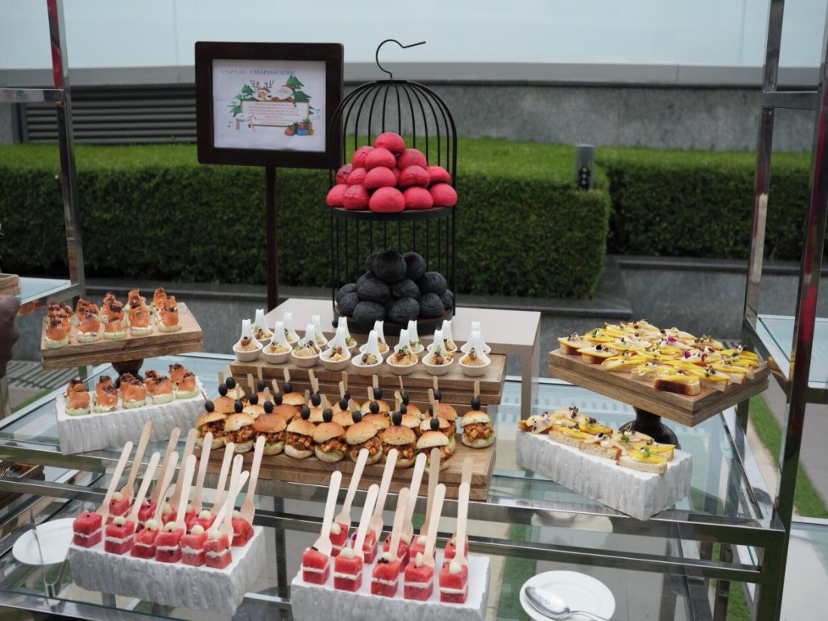 Buffet com diversos alimentos, incluindo petiscos, sanduíches e sobremesas, expostos em mesas de vidro.