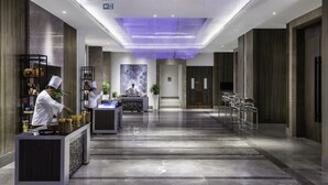 Un vestíbulo de hotel moderno con un mostrador de recepción alargado, un chef preparando comida y un cuadro grande en la pared.