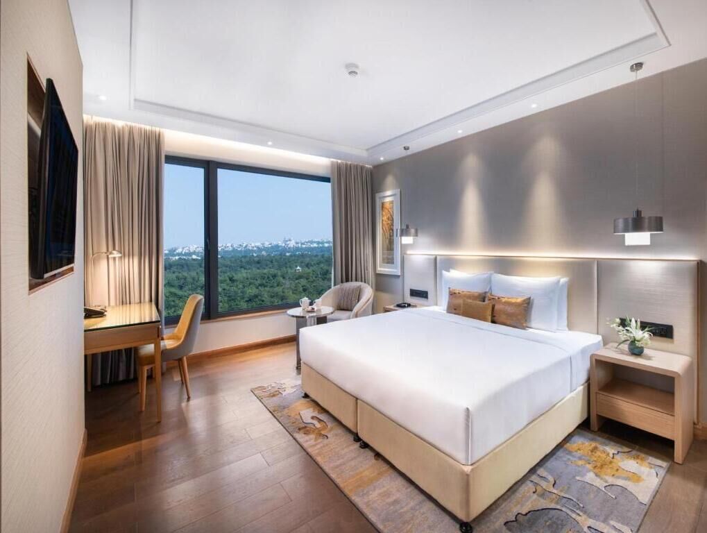 A modern hotel room with a large bed, a desk, a chair, a nightstand, and a view of the outdoors.