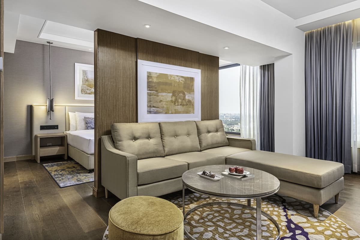 Ett modernt hotellrum med en stor, beige hörnsoffa, ett runt soffbord och en liten pall. Det finns en säng med ett vitt sänggavel, ett nattduksbord med en lampa och ett stort fönster med gardiner.