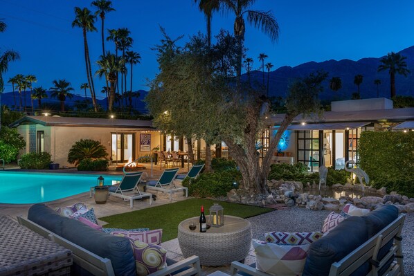 A poolside area with lounge chairs, a dining area, and a view of mountains.