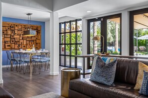 A modern living room with a brown leather sofa, dining area with blue chairs, a wooden wall panel, and large windows.
