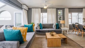 A modern living room with a grey sofa, teal and yellow cushions, a wooden coffee table, and a dining area with wicker chairs. - Seaside Apartment @ Perna Perna Umdloti (eMdloti)